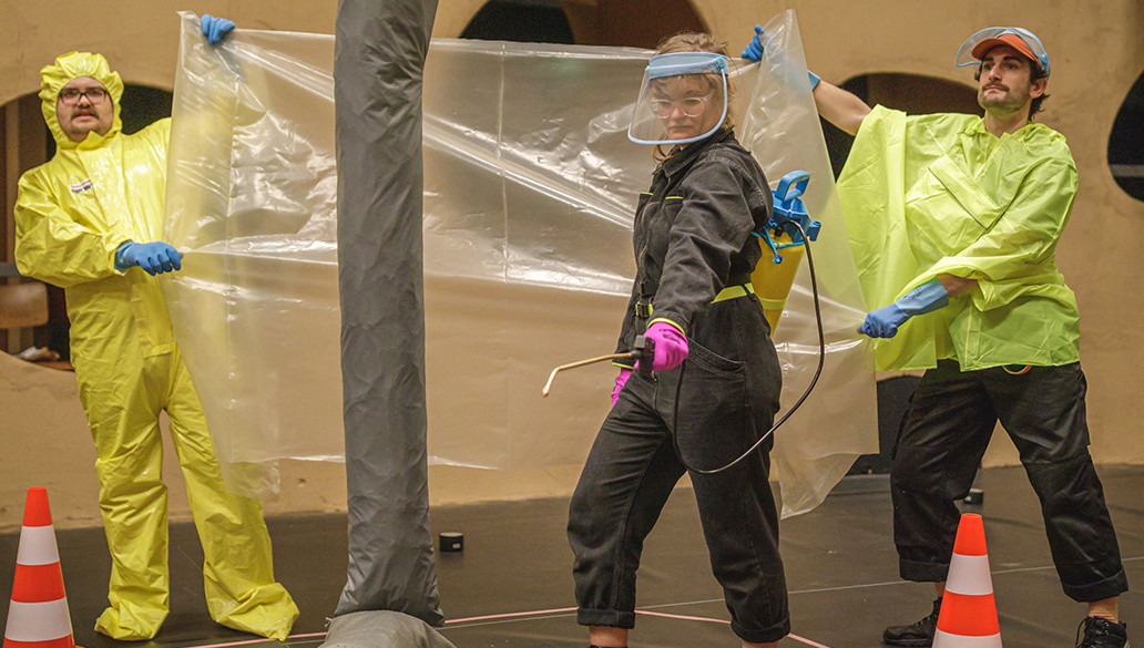 Zwei Personen in Schutzkleidung halten eine Plastikfolie in der Hand, vor ihnen steht eine Person mit einer Art Schlauch in der Hand