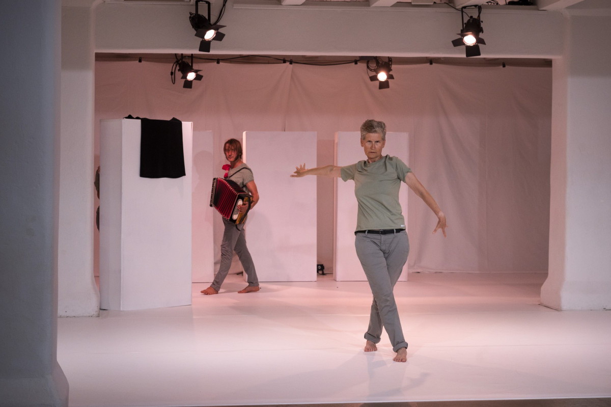 Eine weiße Bühne, im Vordergrund eine Performer*In mit ausgebreiteten Armen, im Hintergrund eine Akteur*In mit Akkordeon